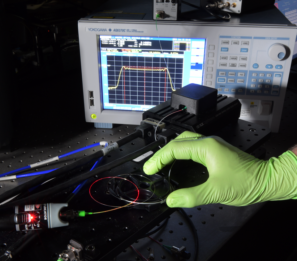 A gloved hand holding an optical fibre carrying red laser light beside laboratory measurement equipment.